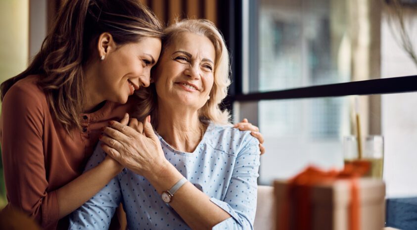 Older mom smiling while side hugging her adult daughter.