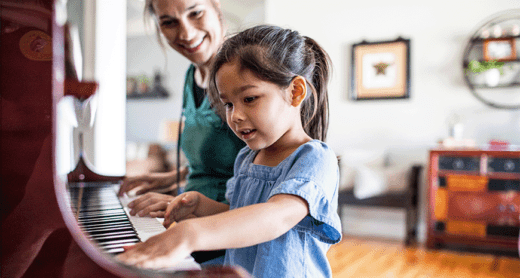 A mother and daughter sitting at a piano in their living room. The mother, while playing, looks over at the daughter who is playing as well.