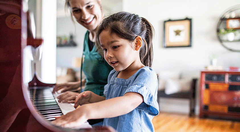 A mother and daughter sitting at a piano in their living room. The mother, while playing, looks over at the daughter who is playing as well.