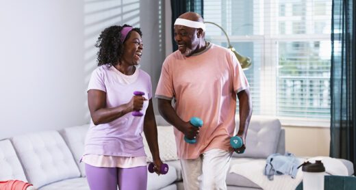 A senior African-American couple exercising together at home in their living room. They are staying fit and active during retirement. The man, in his 70s and his wife, in her 60s, are looking at each other as they do aerobic exercises with hand weights.