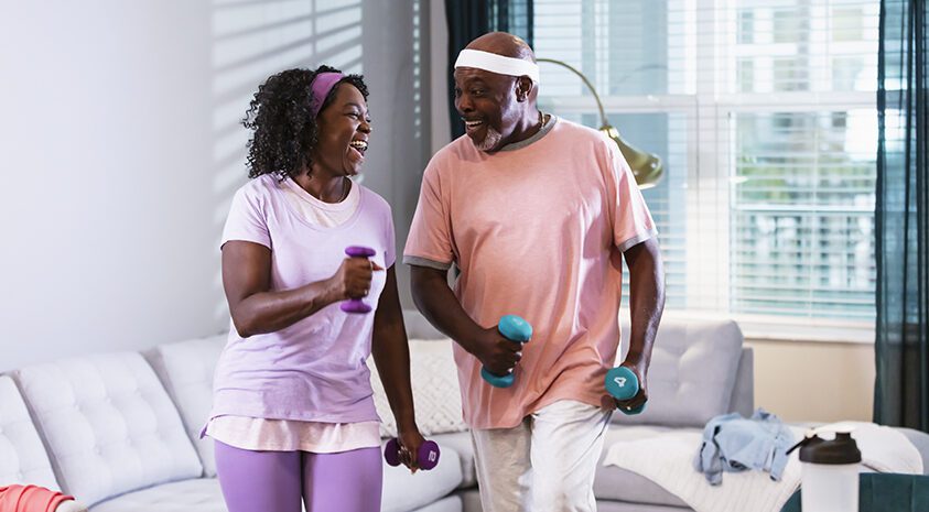 A senior African-American couple exercising together at home in their living room. They are staying fit and active during retirement. The man, in his 70s and his wife, in her 60s, are looking at each other as they do aerobic exercises with hand weights.