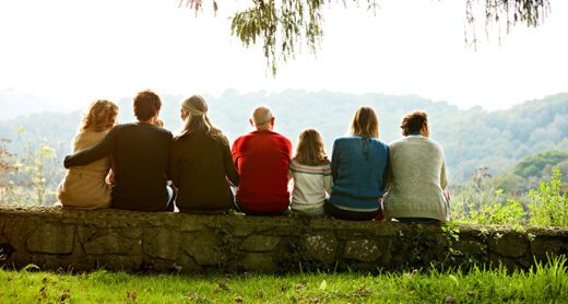 Seven family members sit together on a stone wall looking out at a scenic mountain landscape.