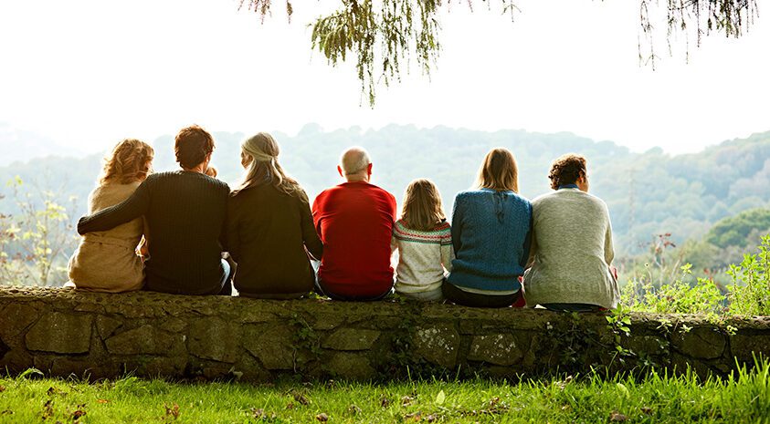 Seven family members sit together on a stone wall looking out at a scenic mountain landscape.