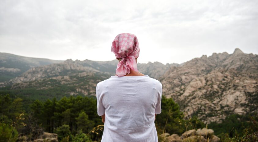Woman with cancer sitting facing a canyon. She wears a pink scarf on her head.