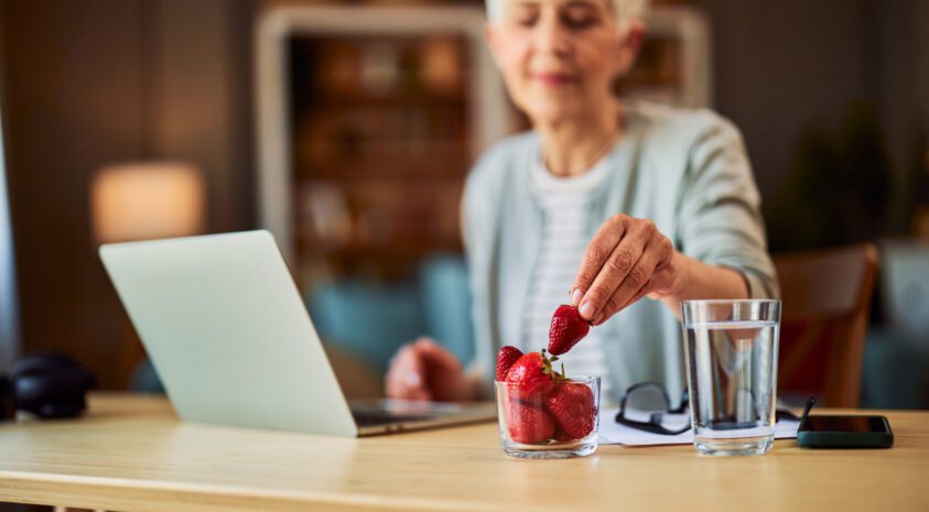 A senior adult woman having a strawberries as a snack while sitting at a desk in front of a laptop.
