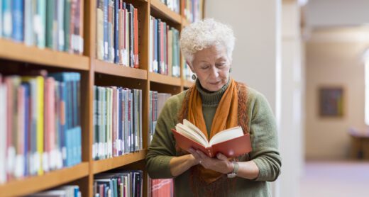 Older woman reading book in library standing next to a bookcase.