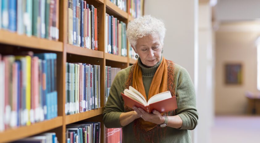 Older woman reading book in library standing next to a bookcase.