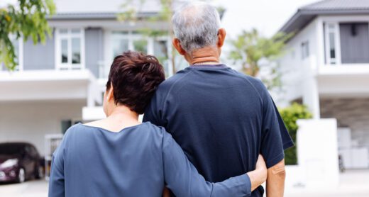 Older couple with woman's arm around man's back shown from behind looking at front of a house.