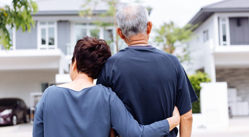 Older couple with woman's arm around man's back shown from behind looking at front of a house.