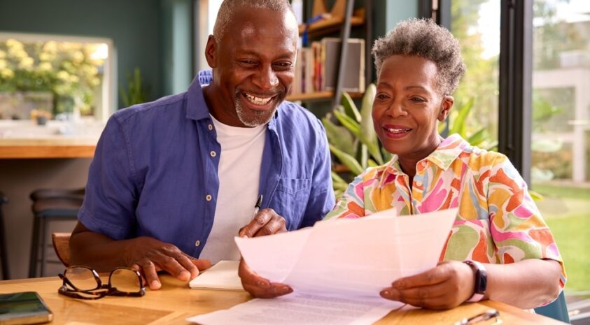 Senior couple smiles while looking at papers at their kitchen table.