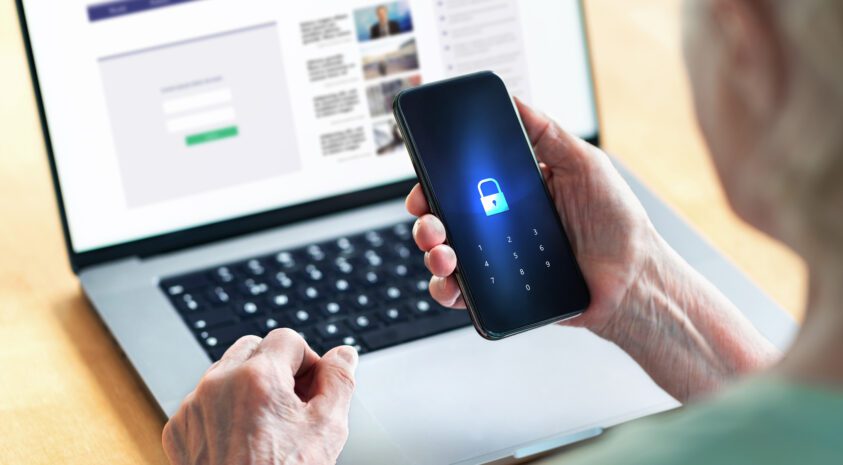 Senior man holds smartphone that's locked while sitting at an open laptop.
