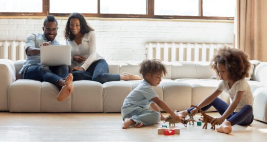 Younger couple sits on their couch researching on computer while smiling as their children play on the floor in front of them.