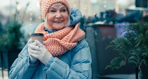 Cheerful older woman spending time outdoors in a cold winter day and smiling while holding a cup of coffee.