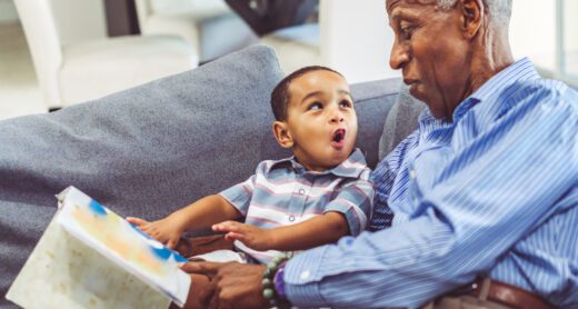 Adorable toddler with his grandfather relaxing at home reading a book together.