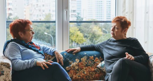 Two mature women in serious conversation while sitting on a couch in front of a window.