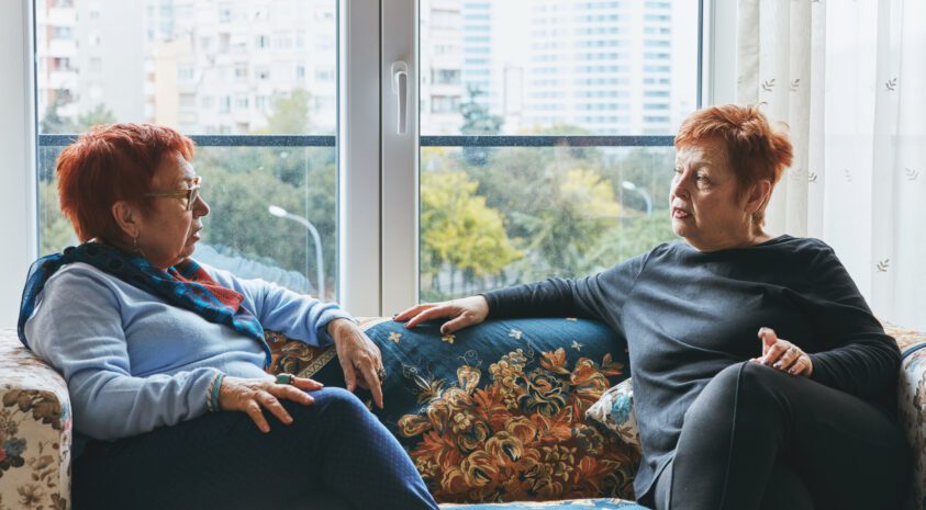Two mature women in serious conversation while sitting on a couch in front of a window.