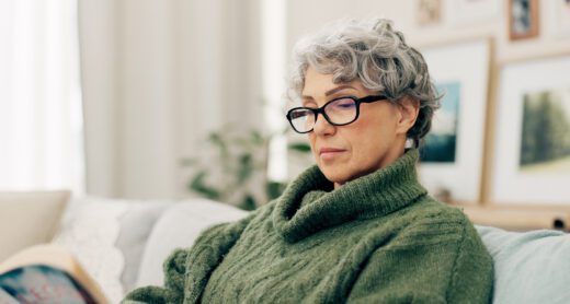 Mature woman with glasses wearing a green sweater reads a book on her couch.