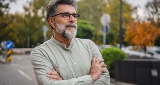 Older man with arms crossed wearing glasses looking into the distance outside.