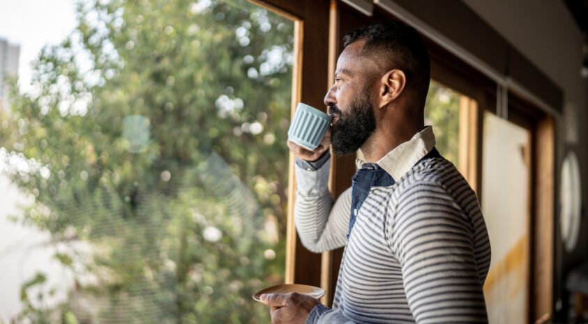 Mature man drinking coffee from a mug while looking out the window of his home.