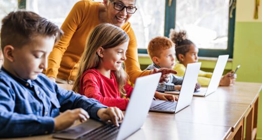 Retired woman smiling leaning down to children in a classroom at a table with open laptops.