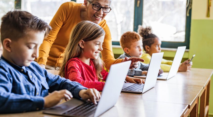 Retired woman smiling leaning down to children in a classroom at a table with open laptops.