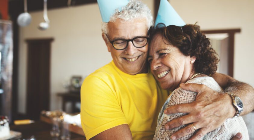 Mature couple wearing birthday party hats embracing and smiling.