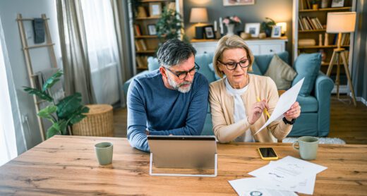 Mature couple sits at kitchen table with an open laptop and papers going over finances.