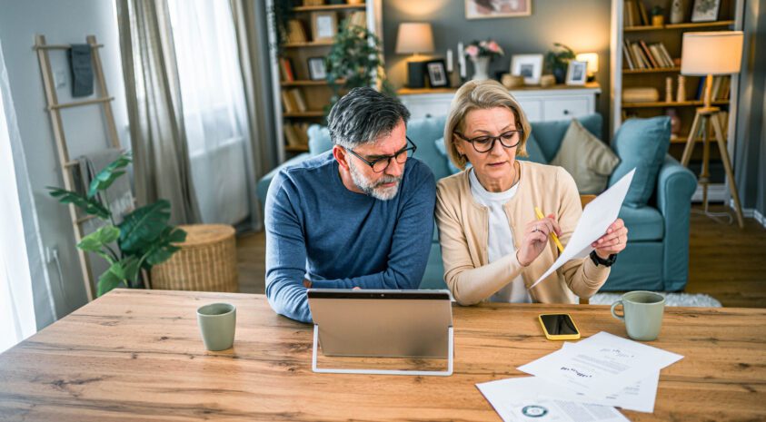 Mature couple sits at kitchen table with an open laptop and papers going over finances.