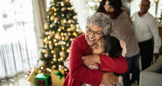 A grandmother hugs her granddaughter at a Christmas celebration at home. A Christmas tree with gifts is lit up in the background.