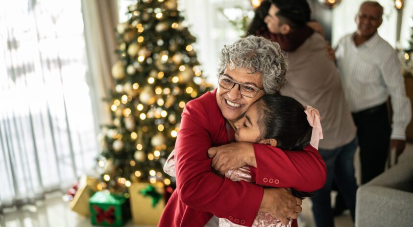 A grandmother hugs her granddaughter at a Christmas celebration at home. A Christmas tree with gifts is lit up in the background.