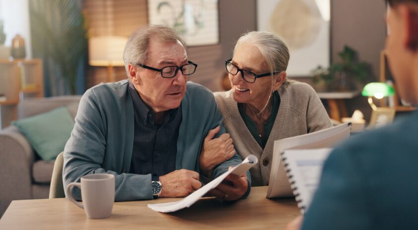 Older couple wearing glasses smiling at each other while going over paperwork with an advisor.