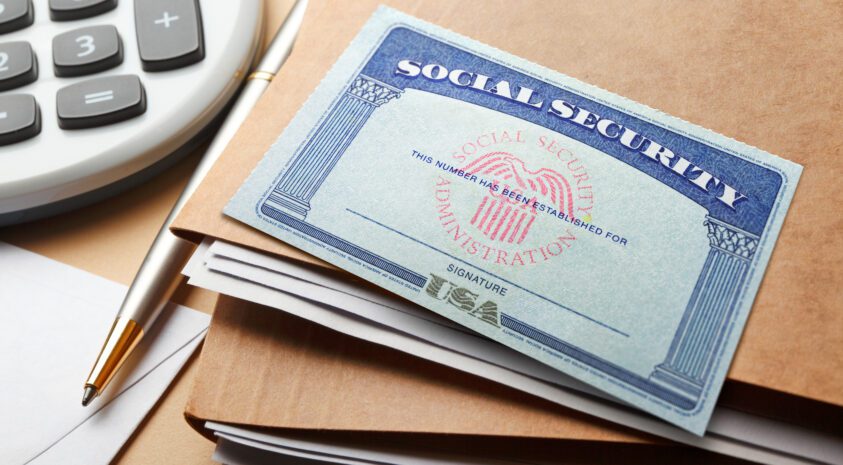 A Social Security card rests on top of a stack of file folders and next to a calculator and a pen.