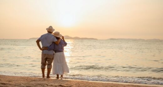 Older couple standing together on a beach at sunset, looking out at the ocean and enjoying a peaceful retirement moment.