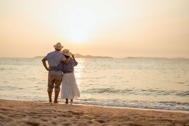 Older couple standing together on a beach at sunset, looking out at the ocean and enjoying a peaceful retirement moment.
