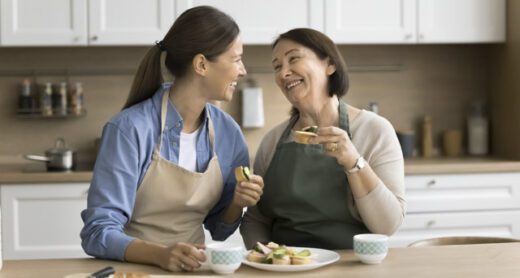 Happy mature mother and adult daughter woman in aprons eating homemade sandwiches in kitchen