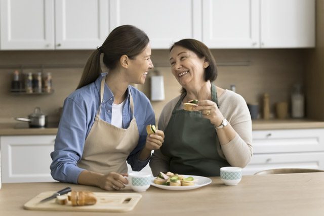 Happy mature mother and adult daughter woman in aprons eating homemade sandwiches in kitchen
