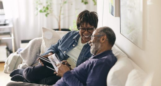 Happy senior woman with digital tablet talking to man sitting on sofa at home